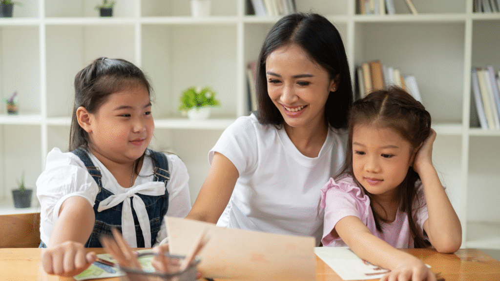 Seorang babysitter sedang mendampingi dua anak kecil saat belajar dan menggambar di meja.